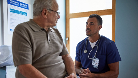 Image shows Hispanic healthcare professional speaking to older Hispanic man in healthcare setting.