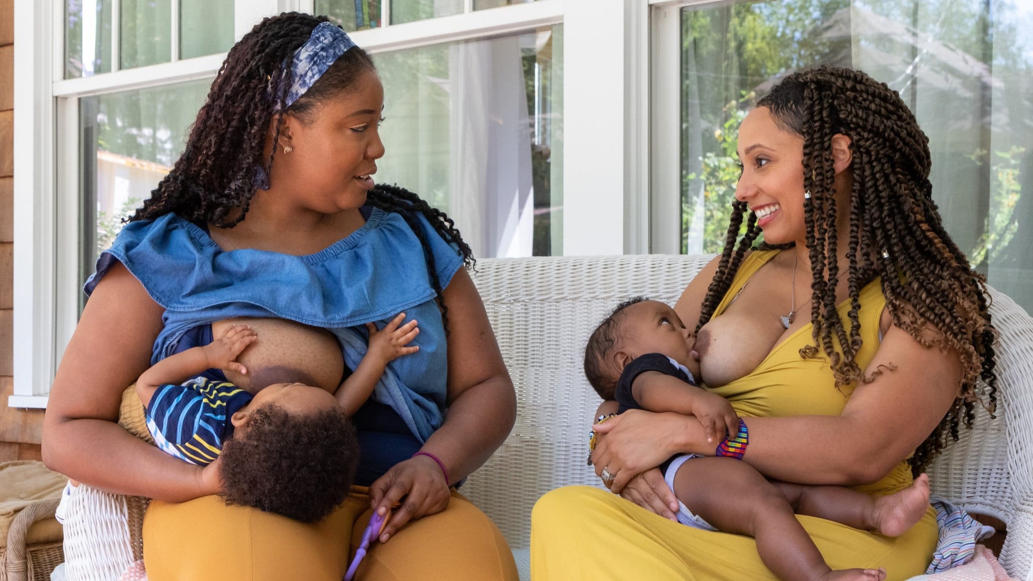 Two women breastfeed two children while sitting on a porch outside a house.