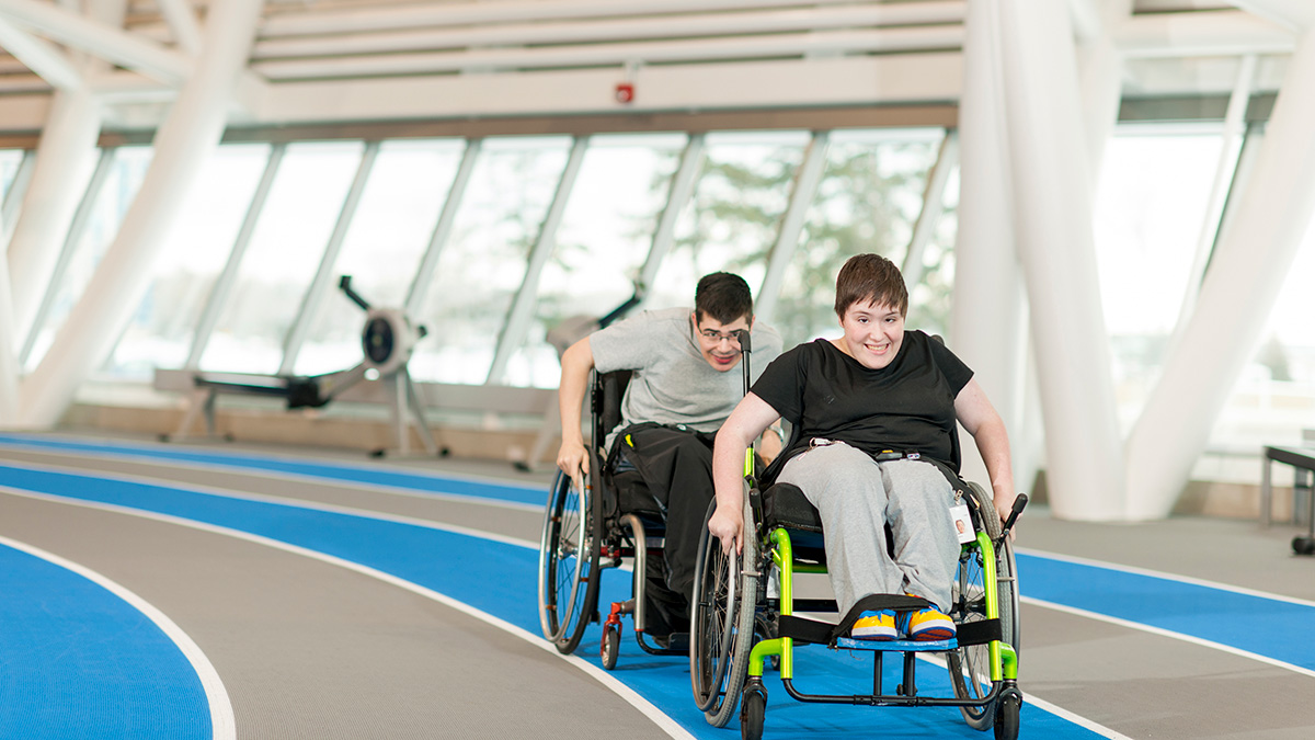 Two people racing in wheelchairs on an indoor track. The person in a black shirt is ahead of the person in a grey shirt.