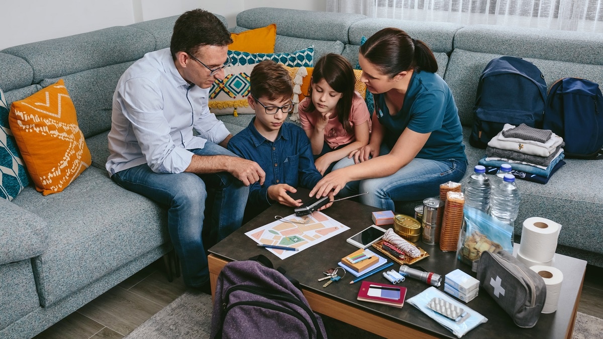 A family sits on a couch around a table with emergency supplies and a map. Adults and children are focusing on how to use an emergency radio.