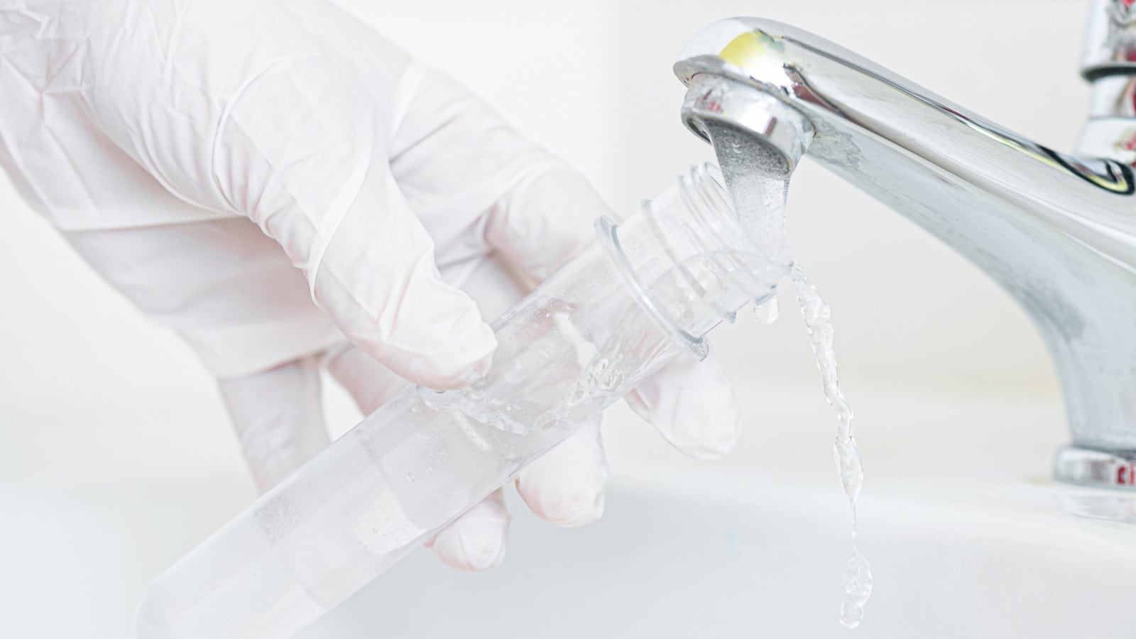 Gloved hand holding a test tube under a running faucet to collet water