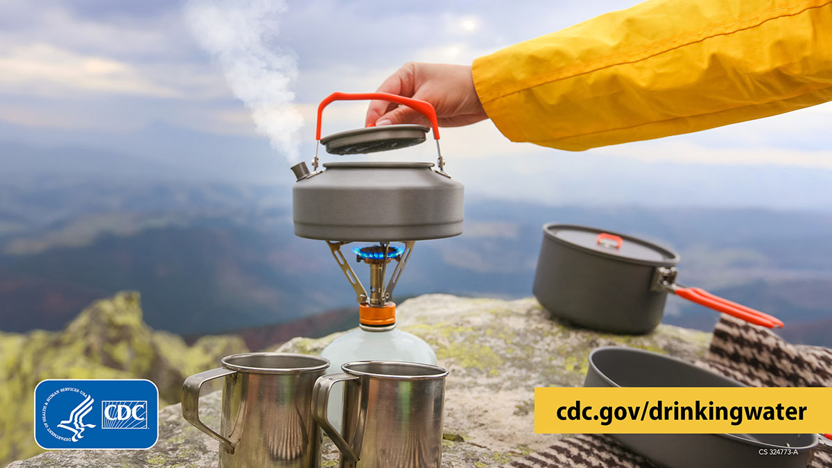 Hiker on a peak overlooking mountain ranges. Hiker lifts the lid of a kettle of water boiling on a portable stove, with pots and mugs nearby.
