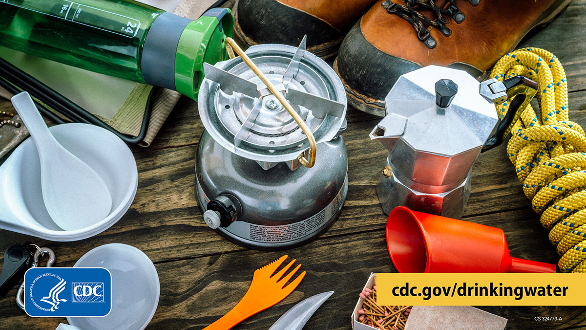Table with camping supplies on it, including a portable gas stove, water kettle, water bottle, matches, hiking boots, bowls, and utensils.