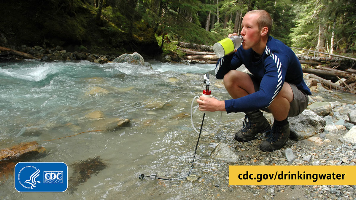 Hiker holding a water filter in a mountain stream in one hand and drinking out of a water bottle with the other hand.