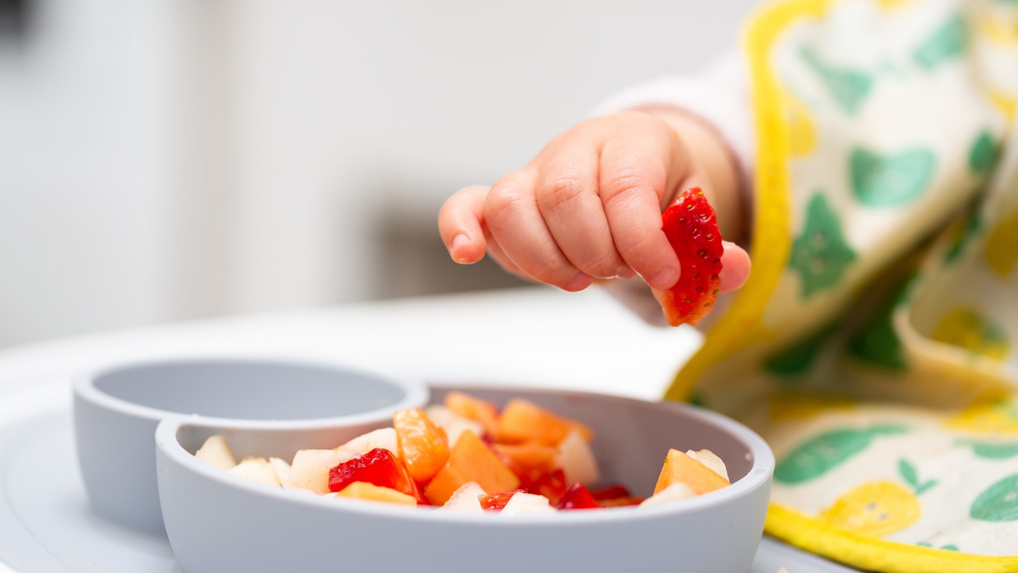 Close up of baby hand with a small piece of fruit over a plate of diced fruit