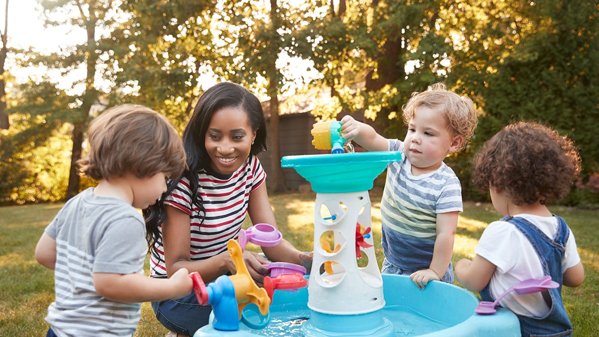 Teacher kneels down to supervise young children while they play with water outdoors.