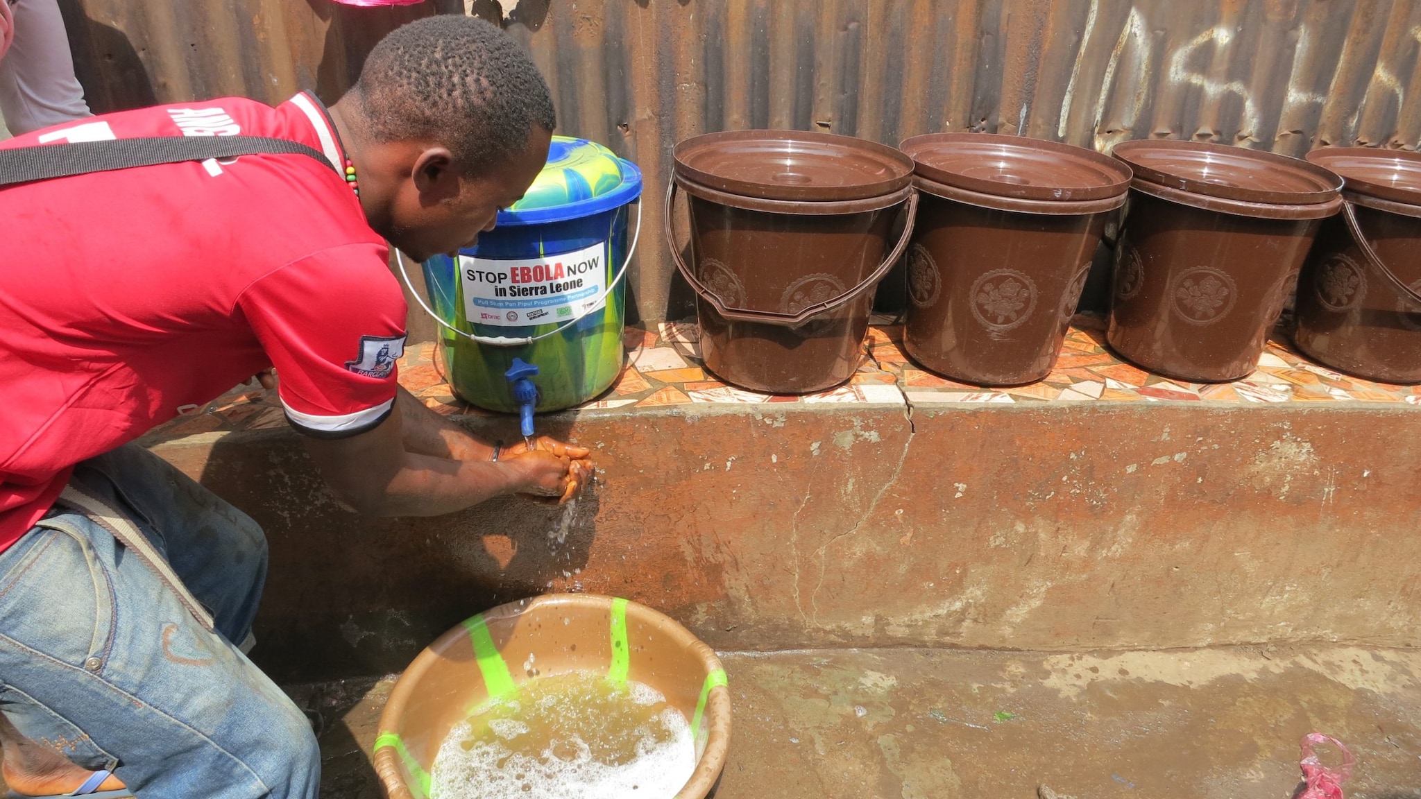 A young man washes his hands using a bucket handwashing station.