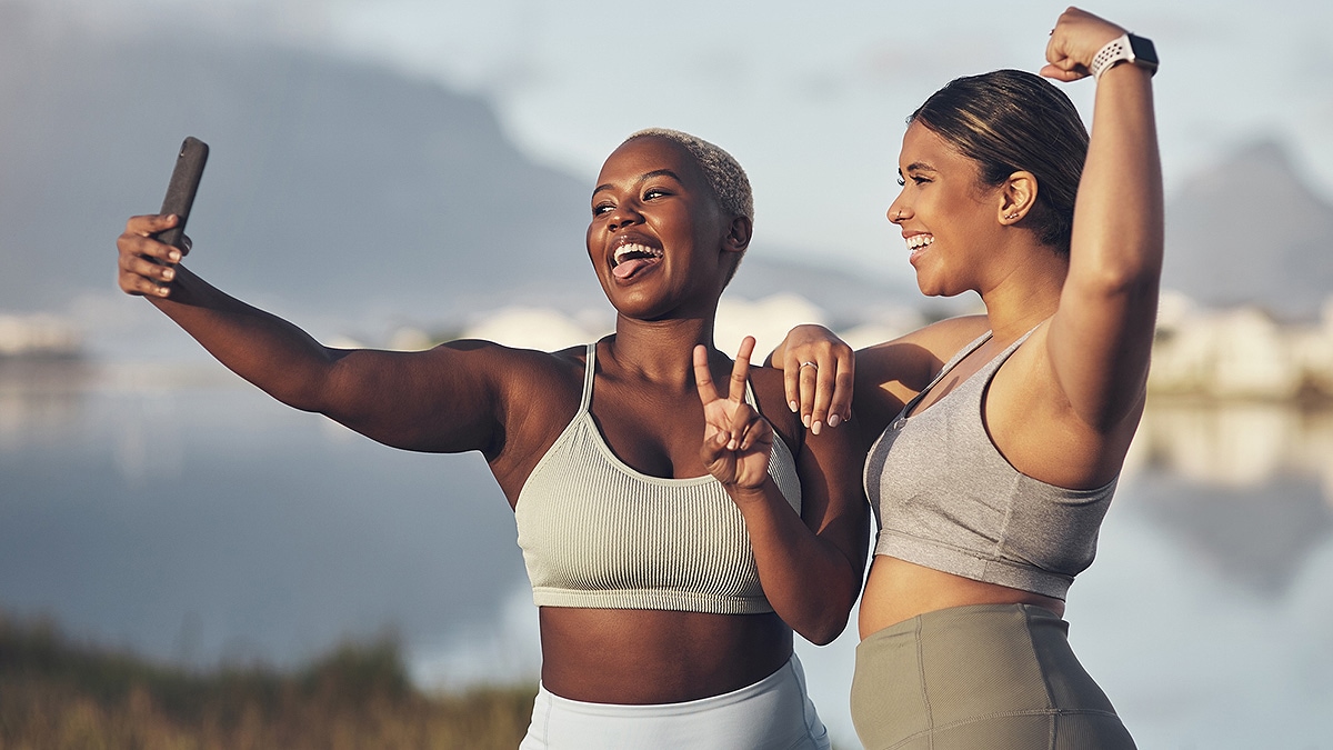 An African-American and a Latina woman posing with a peace sign and flexed bicep for a happy selfie.