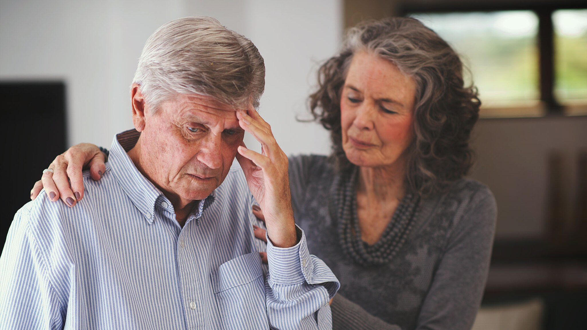 An older woman comforts an older man who is holding his head.