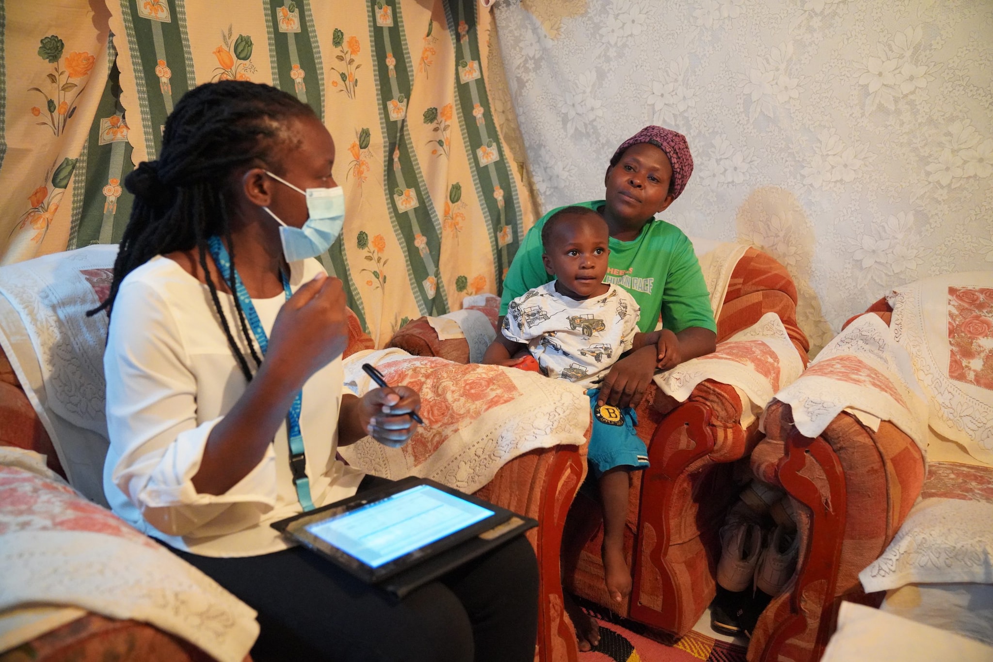 Public health worker interviews mother and child in a living room in Kenya and documents information on a tablet