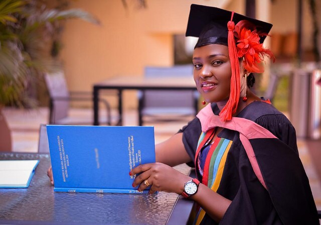 A woman wearing a black cap and gown with red accents is smiling while holding a blue book.