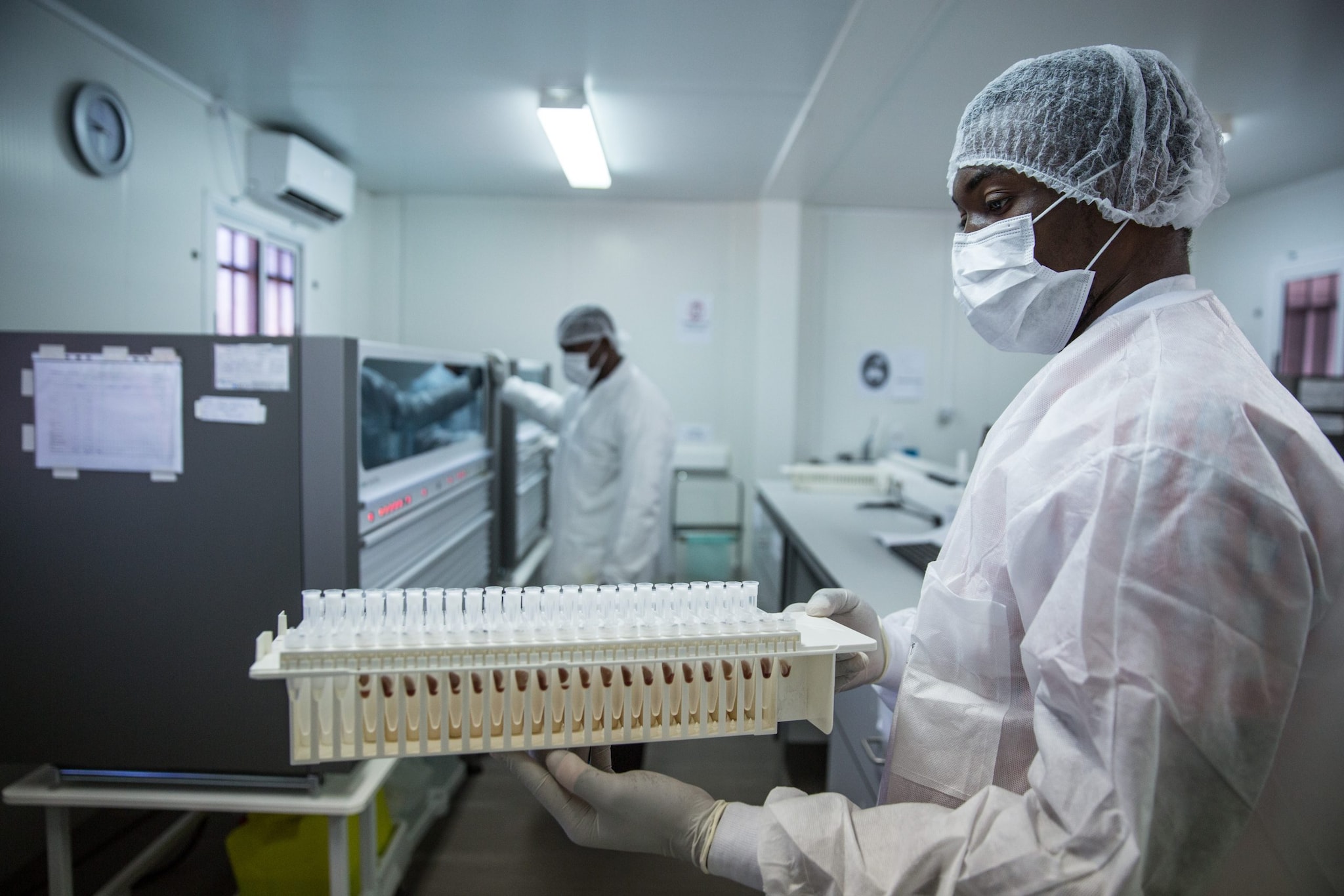 A man wearing white lab gear, blue gloves, a mask, and hair protection stands in a laboratory.