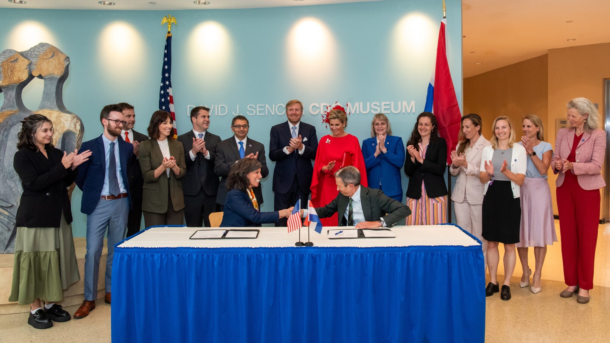 A group of individuals standing and clapping around a table where Dr. Laserson and Prof. Hans Brug are sitting and shaking hands