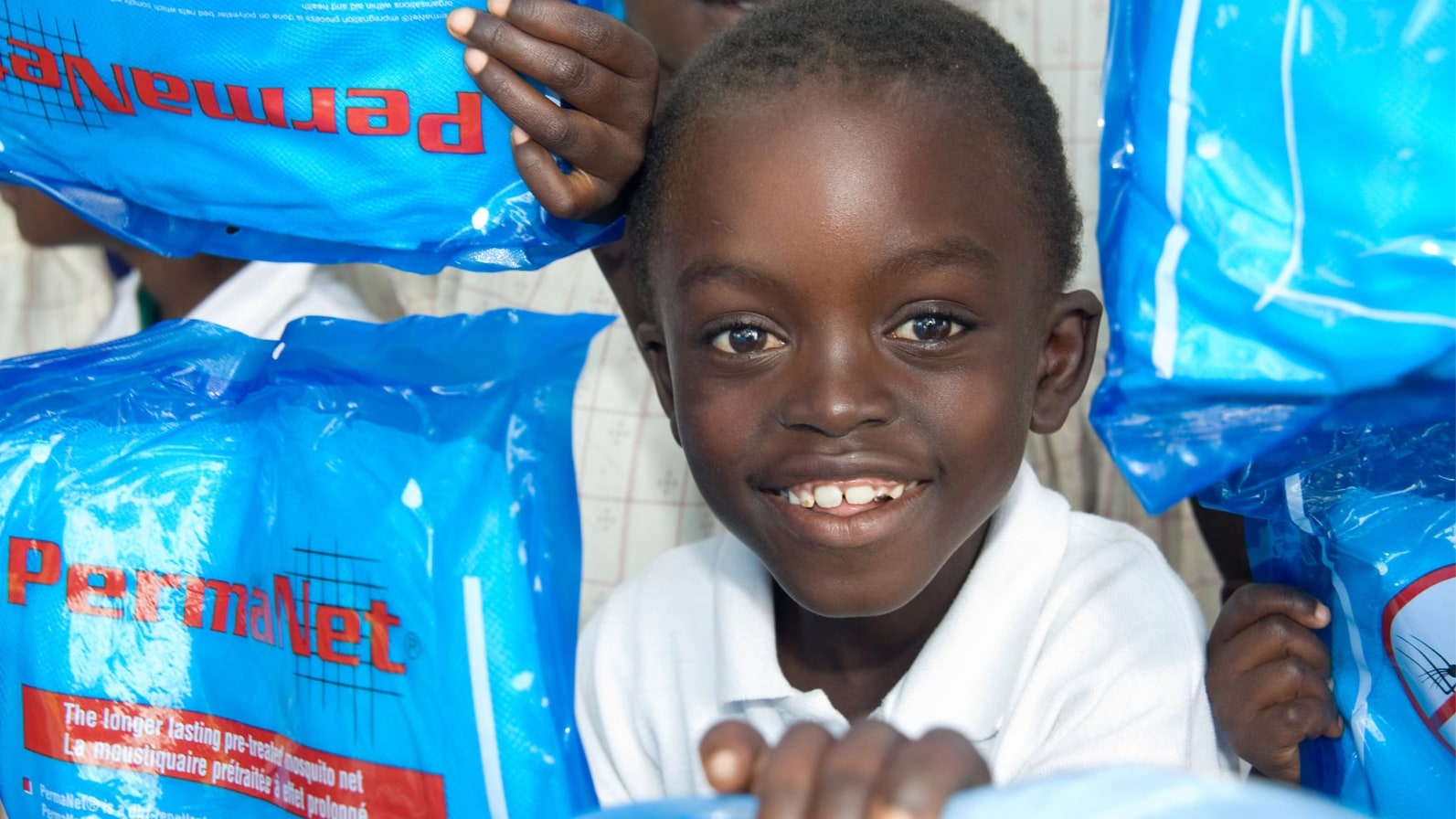 Photo of a young child smiling while people hold up packages of wrapped bed nets around him.