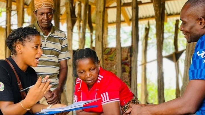 A local community mobilizer in a black shirt sits with three other people, one in a brown stripe shirt, one in a red jersey, and one in a blue shirt, as she talks with them to collect data.