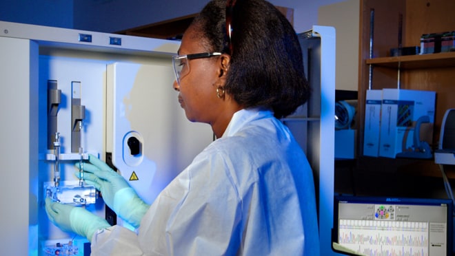 A woman in a lab coat works at a machine in safety glasses.