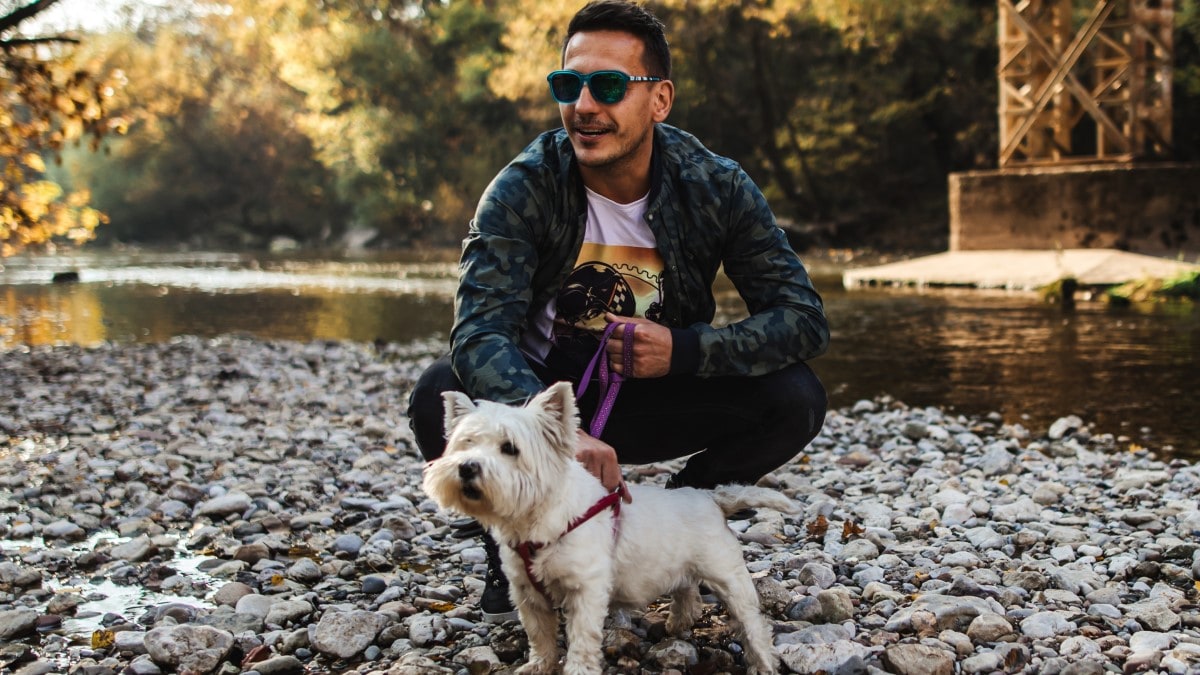 Persona con gafas de sol sosteniendo la correa de un perro pequeño en una costa rocosa con un río en el fondo