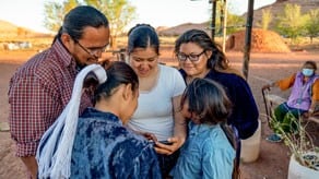 A group of five people in a rural environment standing in a circle looking at a phone together.