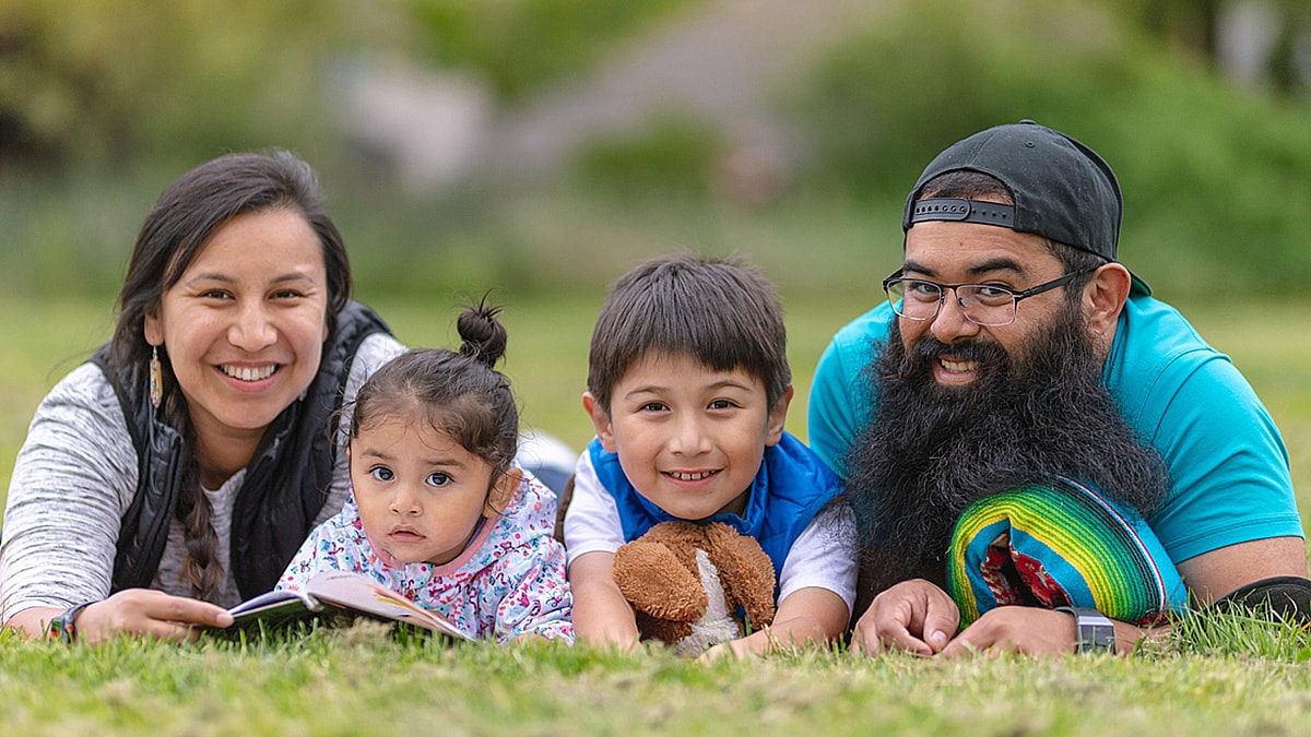 Young, American Indian couple lying in the grass in a park, reading to their two young children.