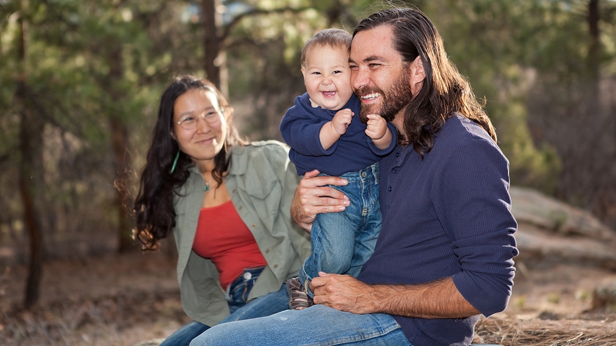 Young American Indian family with mom, dad, and toddler sitting on a rock in the woods.