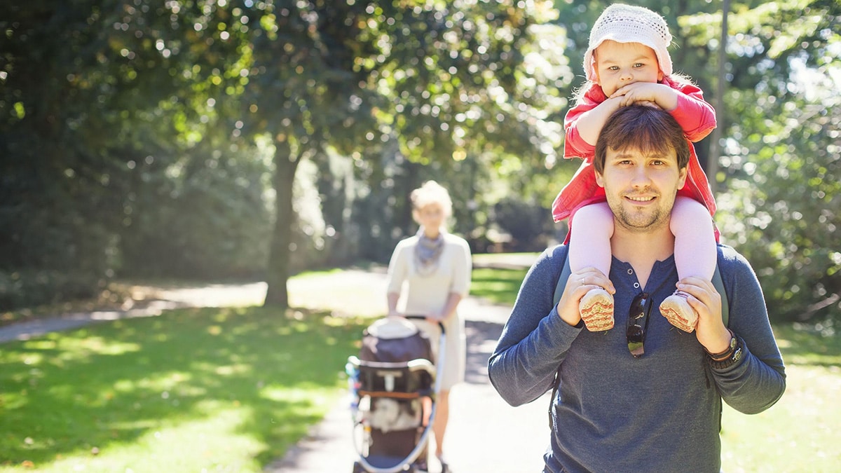 Man walking in the park with toddler on his shoulders, and a woman behind him walking with a stroller