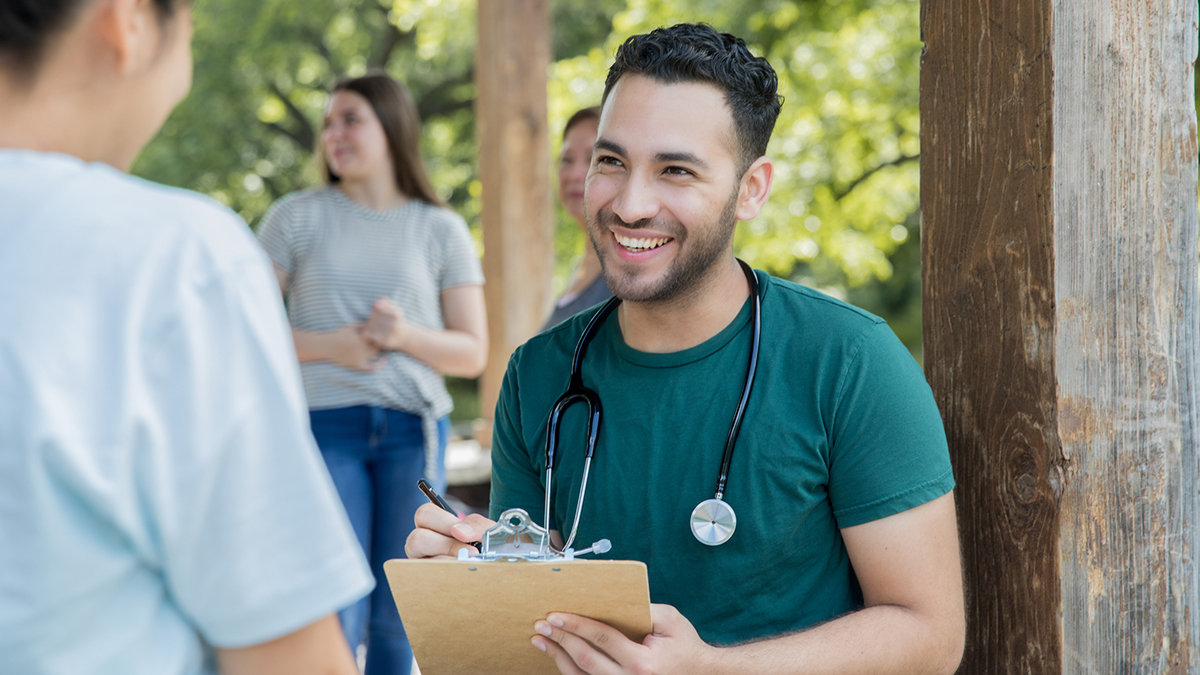 A patient speaking with a doctor outdoors while the doctor takes notes on a clipboard