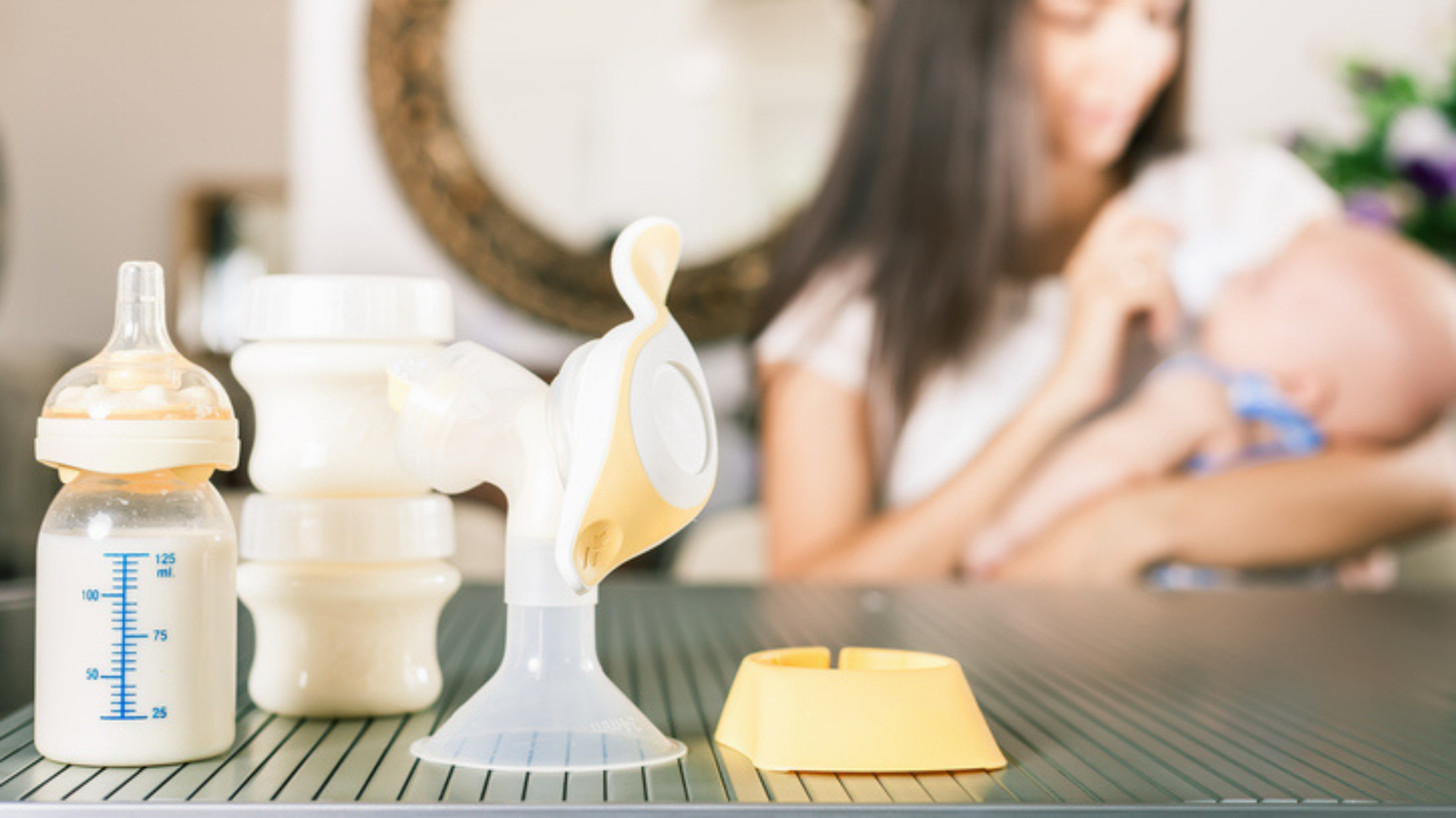 Breast pump parts on a table in front of a woman feeding her infant