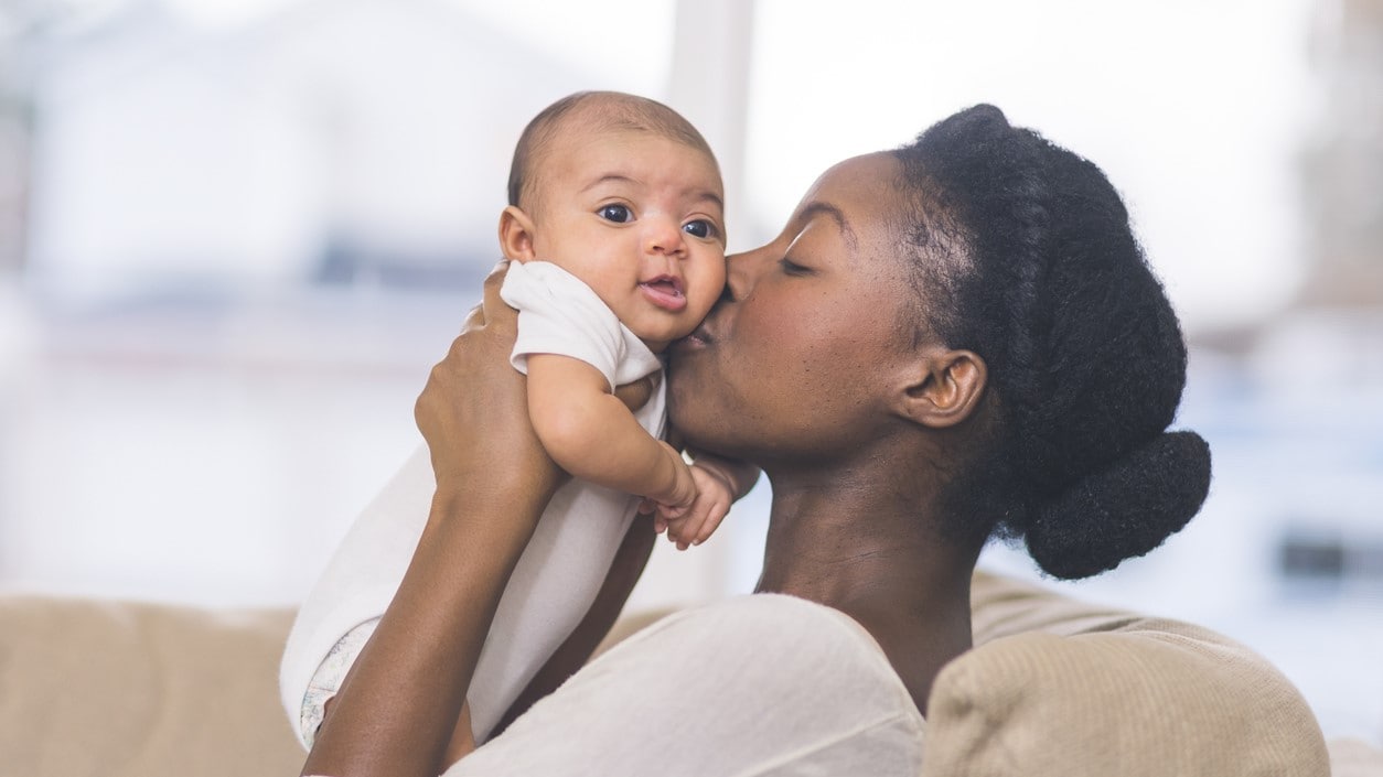 Mother holds her baby up in the air with both hands and kisses its cheek.