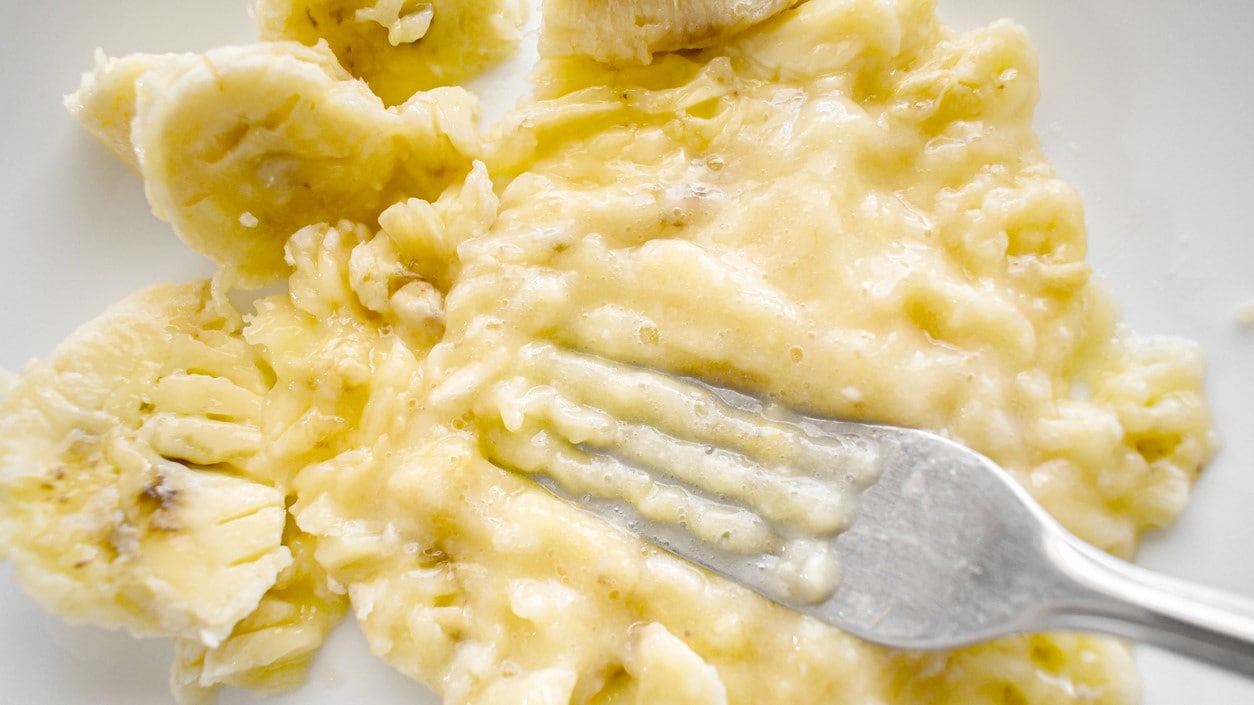 Ripe banana being mashed with a fork on a white plate.