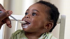 6 month old baby sitting in high chair and being fed solid foods for the first time from a spoon.