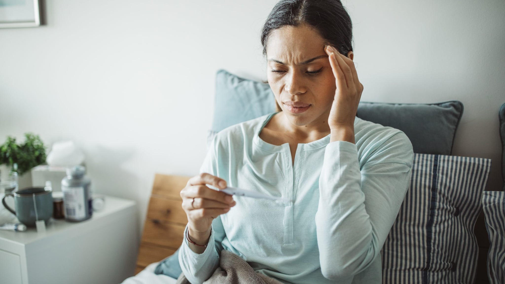 Woman in bed looking at a thermometer and her hand is on her forehead