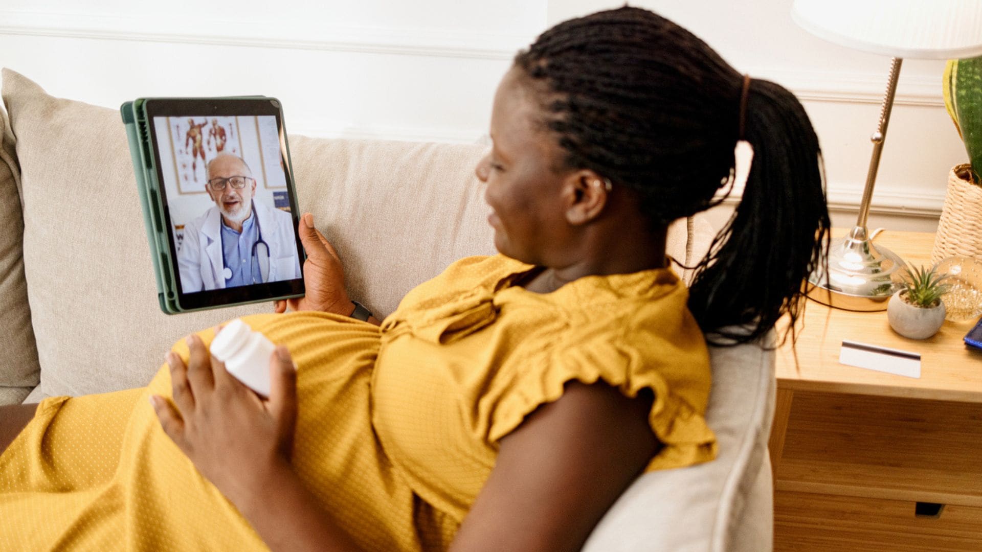 Pregnant woman sitting on a couch with a bottle of medicine talking with her doctor via a tablet.