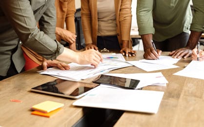 Several people standing around a table covered with papers and computer tablets.