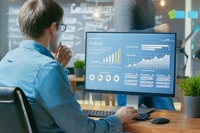 Analyst Works on a Personal Computer Showing Statistics, Graphs and Charts. In the Background His Coworker and Creative Office.