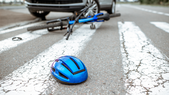 A bicycle lying in a crosswalk with a biker’s helmet lying nearby and a car bumper in the background.
