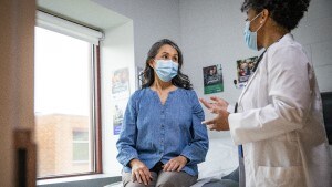 older woman sits in exam room wearing mask and talking with doctor in mask