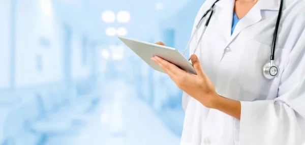 A doctor in a white coat with a stethoscope around their neck uses a tablet to review a report in a hospital hallway.