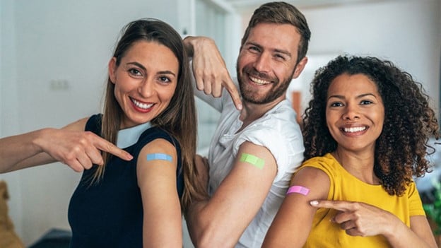 Three people smile and point to their upper arms, showing their colored band-aids.