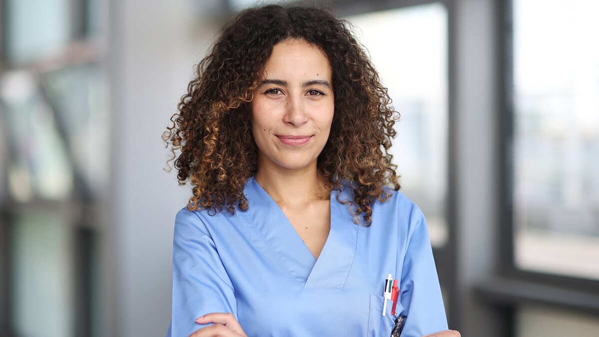 Healthcare worker in scrubs stands with arms crossed looking confident.