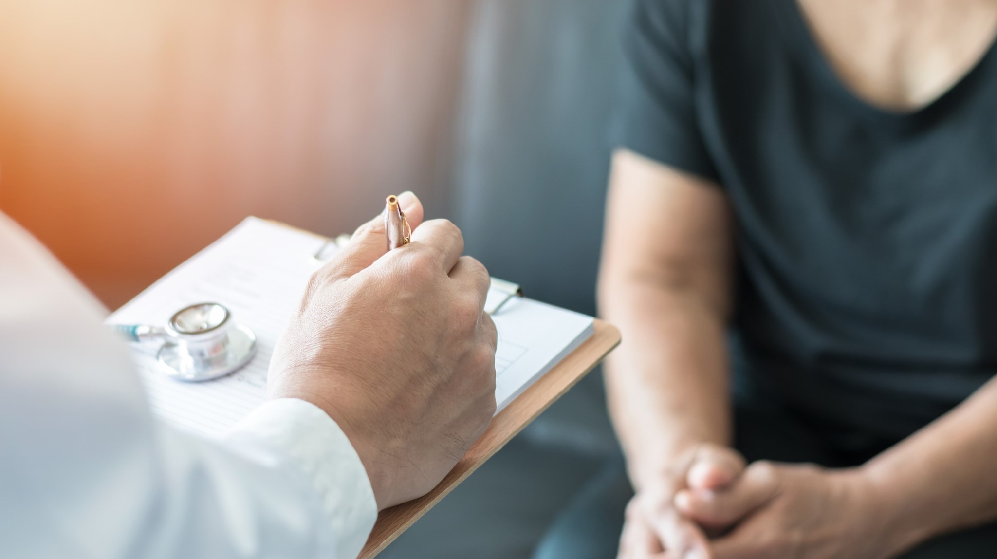 Clinician hands writing on a clipboard standing in front of someone sitting down