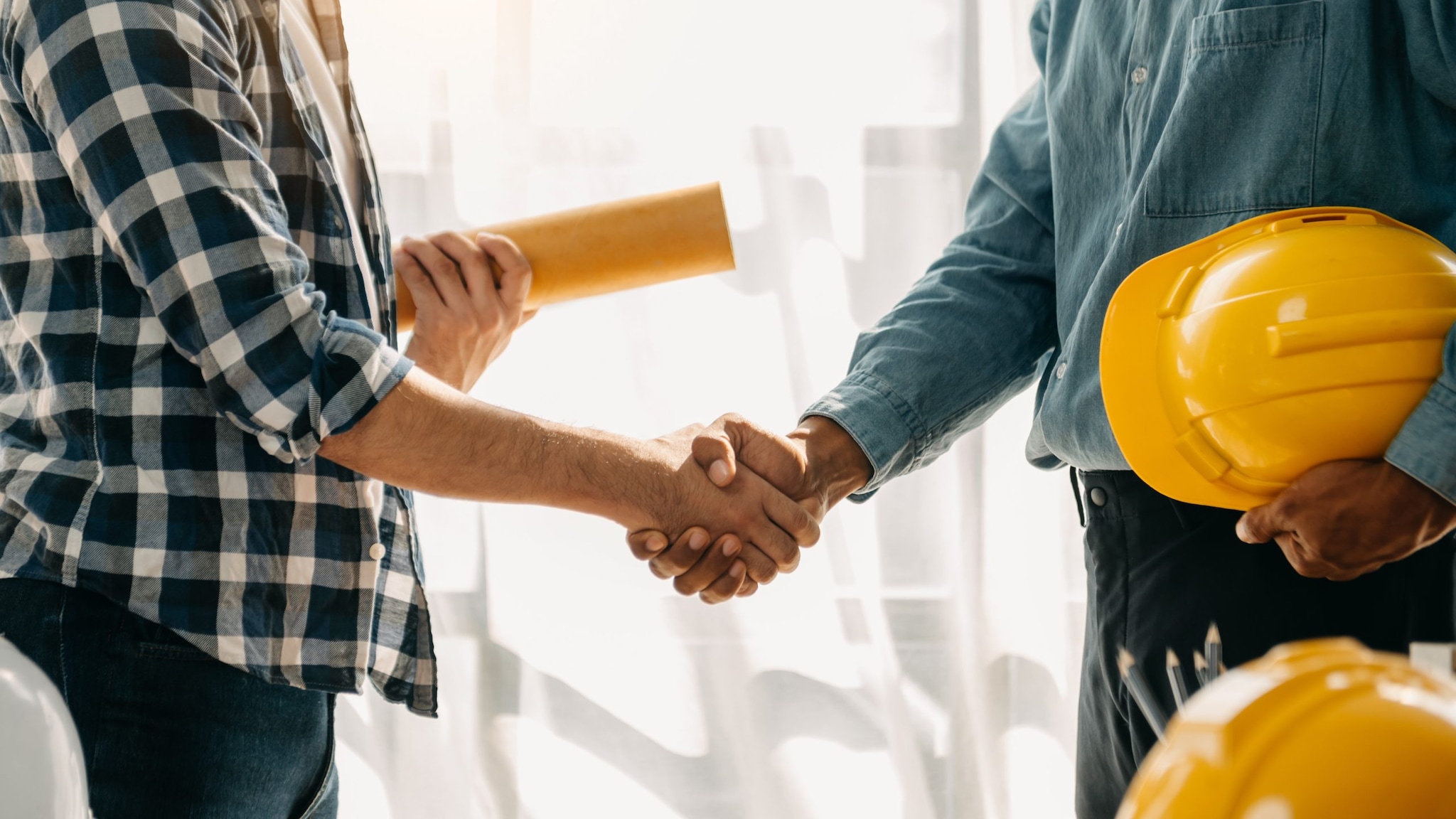 Two people shaking plans, one holds rolled up project plans and the other holds a safety helmet.