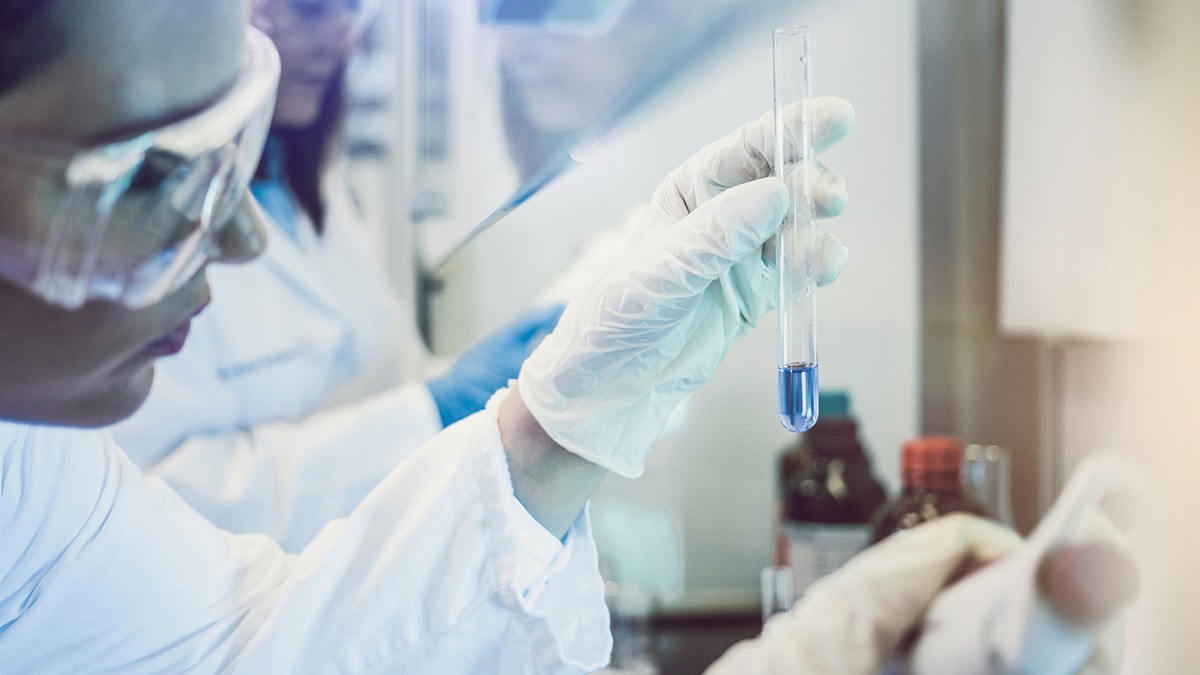 Scientist wearing gloved and eye protection, looking at a test tube.