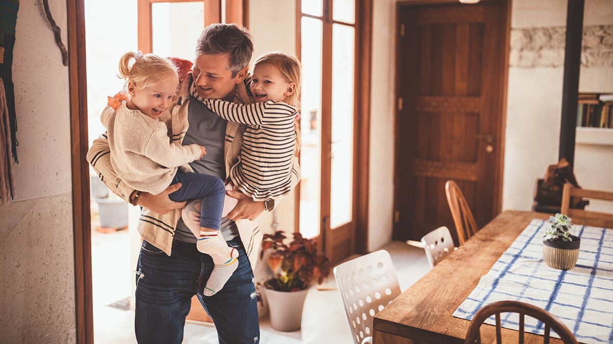 A father has returned home from work and is holding 2 children.