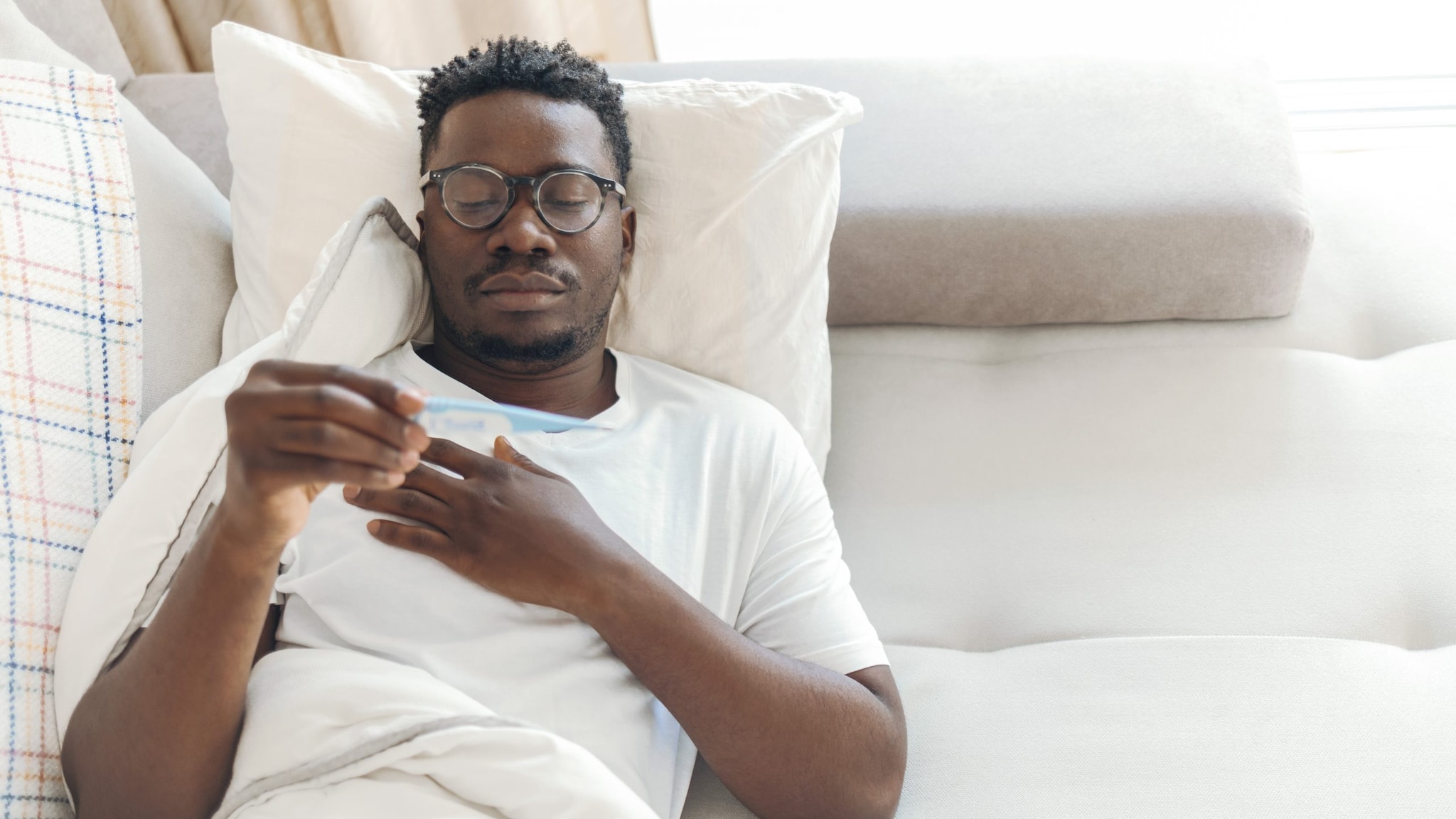 African American man laying on a white couch, taking his temperature.