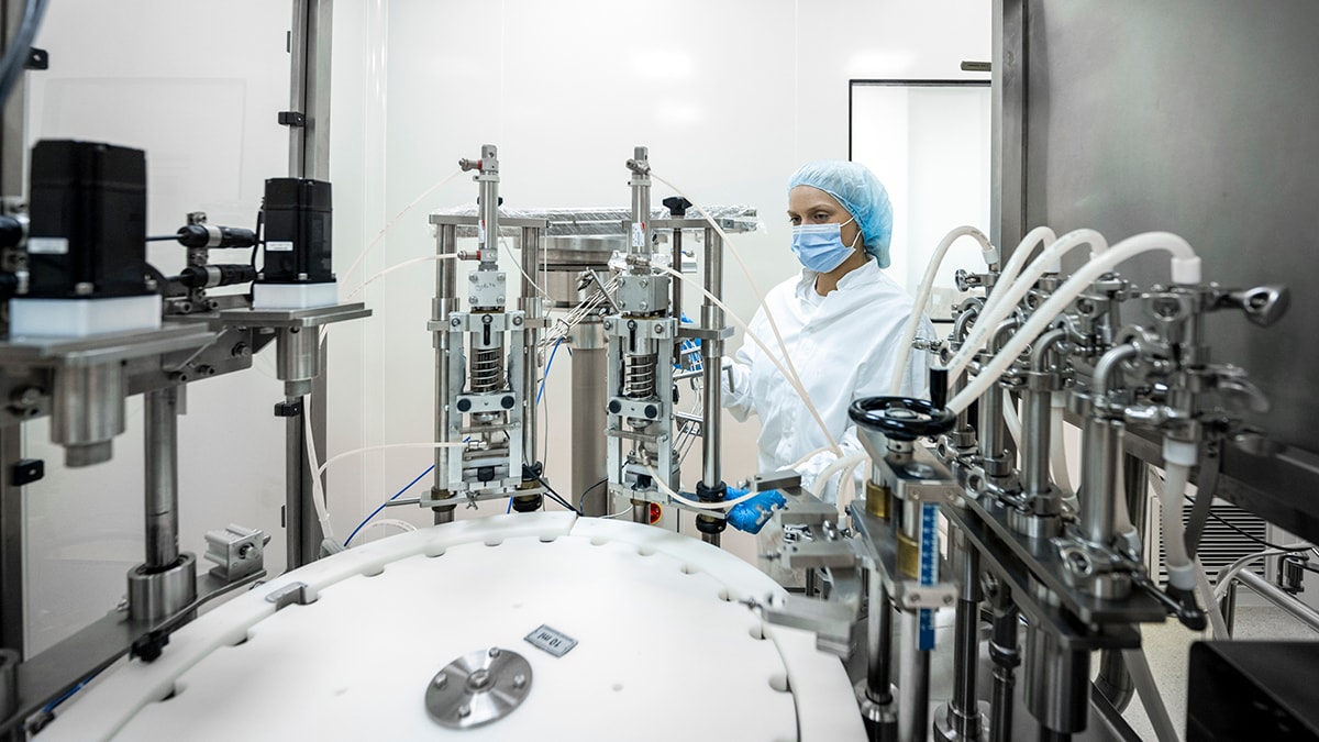 Small brown bottles seen in a laboratory machine during manufacturing in a pharmaceutical factory while a female employee in a protective clothing is controlling the process.