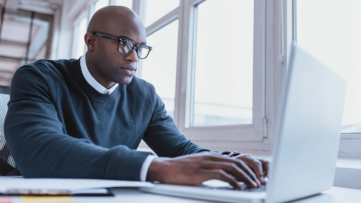 Man wearing glasses and a sweater, working on a laptop at a desk near a window.