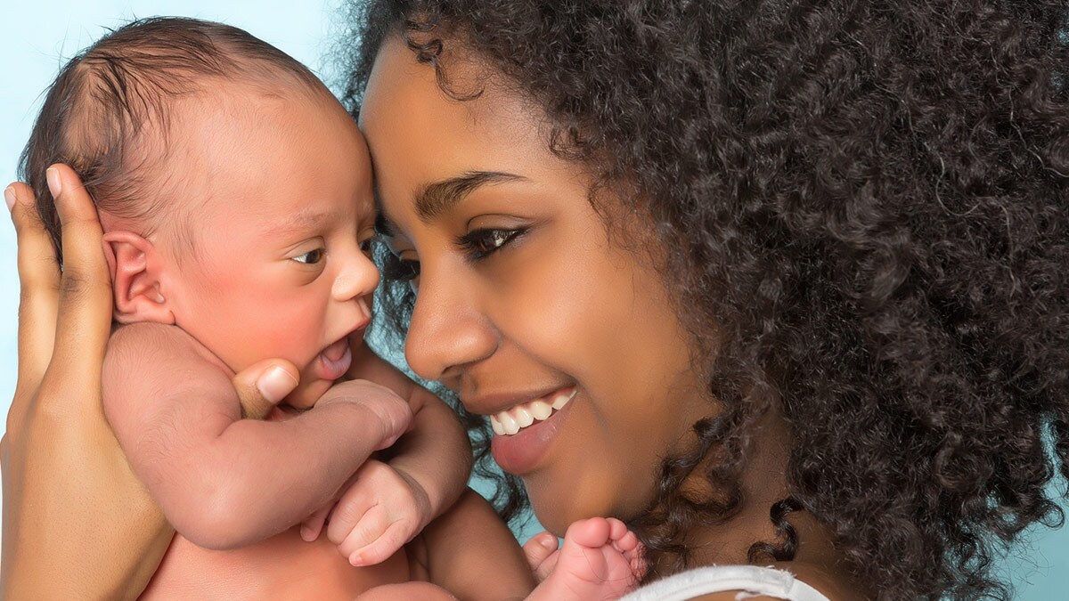 A smiling mother with curly hair lovingly holds her newborn baby, both facing each other closely.