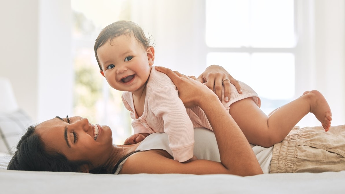 A smiling mother holds her baby while lying in bed.