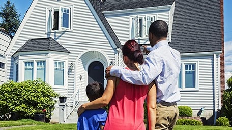 A family with their backs turned stands in front of a house on a sunny day.