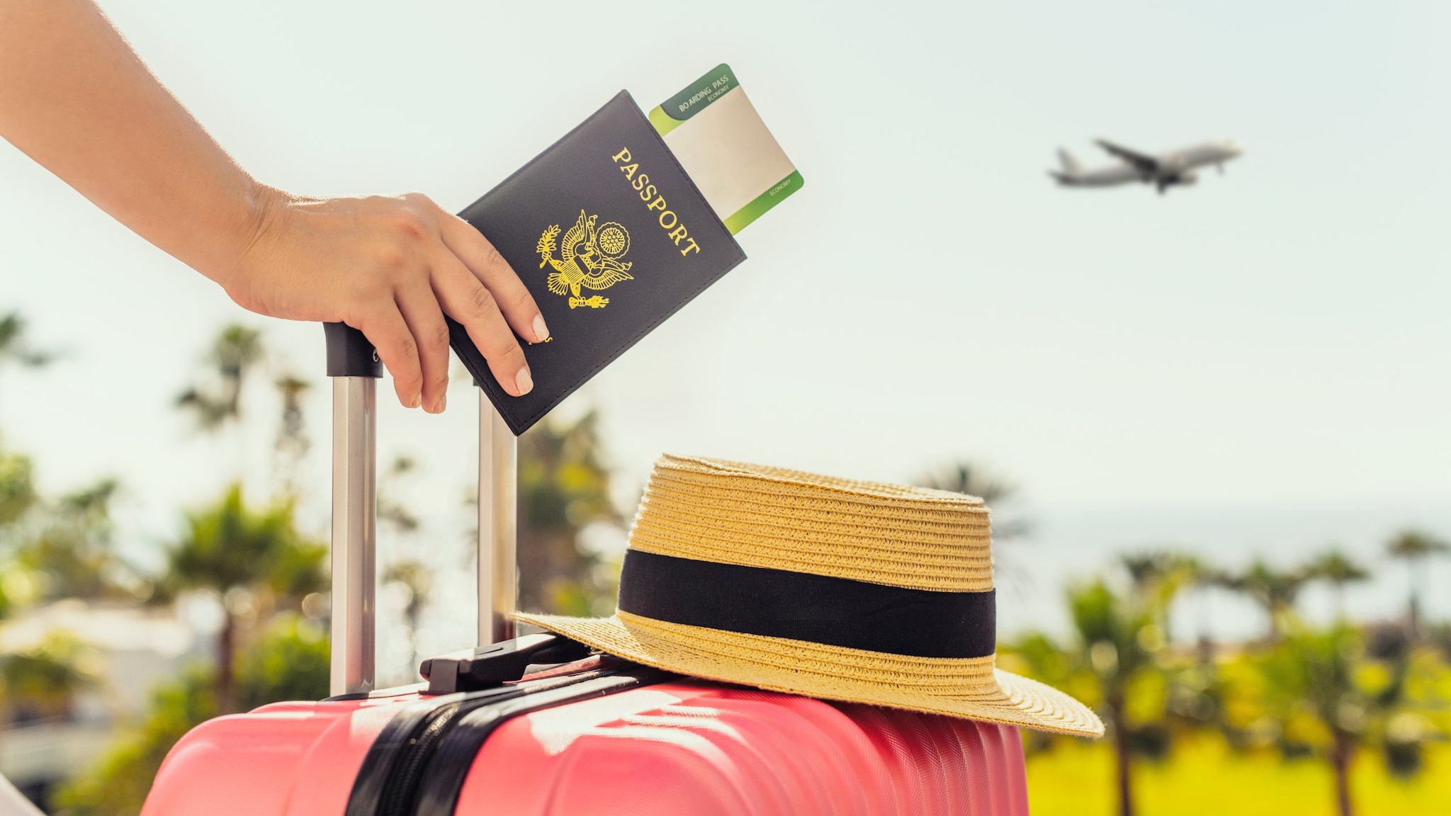 A hand holding a passport and pink suitcase with a hat on top. The person is standing outside watching a plane in the sky.